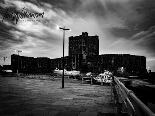 Historic castle silhouette against a dramatic sky, with boats and a waterfront walkway.