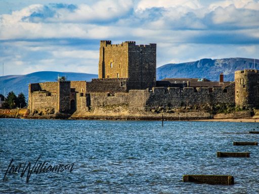 Historic castle on a coastline with mountains in the background and clear blue skies.