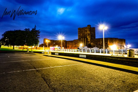 A castle illuminated at night, with a cloudy blue sky and a foreground of a dark parking lot.