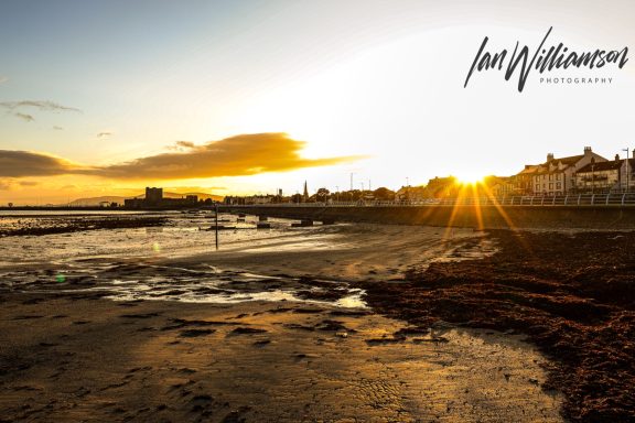 Sunset over a beach, with silhouettes of buildings and tidal pools in the foreground.