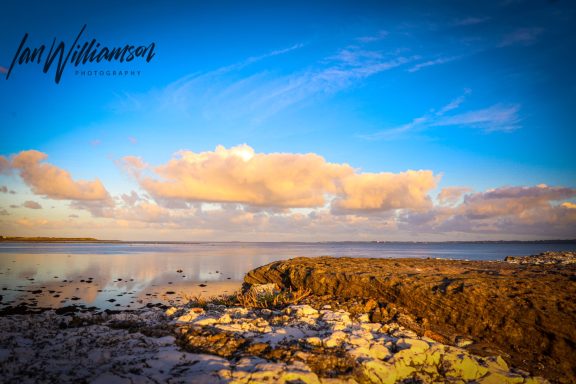 A serene coastal landscape with blue skies, clouds, and calm waters reflecting the scenery.