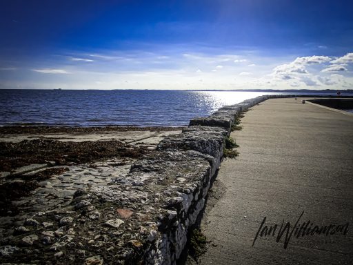 Coastal pathway alongside calm water under a blue sky with scattered clouds.